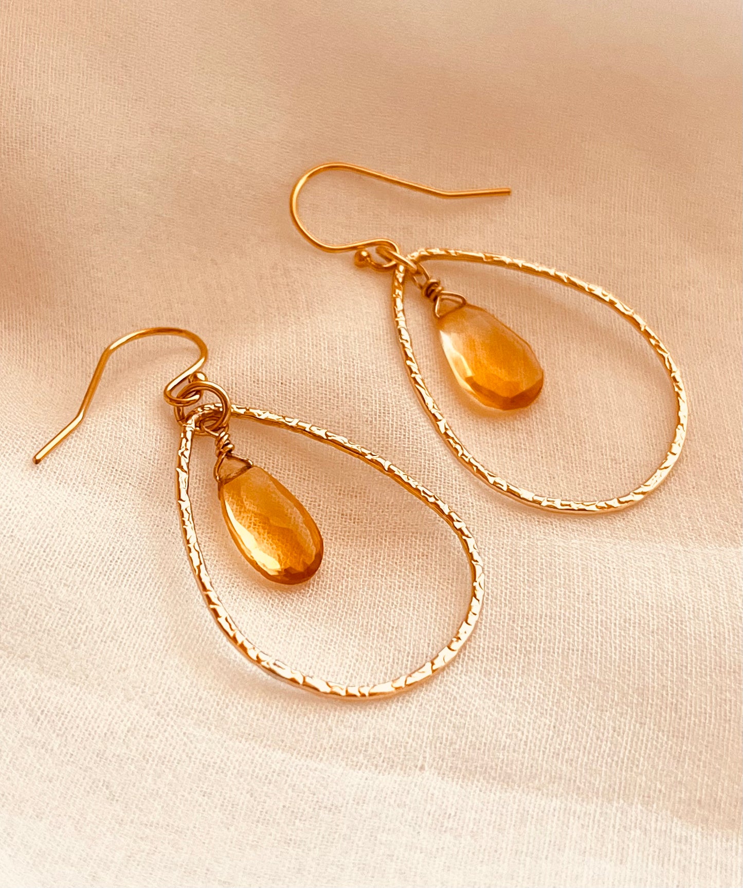 Elegant and lightweight, these handcrafted earrings feature warm yellow teardrop gemstones suspended inside textured 14k gold-filled teardrop hoops. Each stone catches the light with a soft shimmer, adding a refined pop of color to any outfit. The delicate hammered finish on the hoops adds subtle detail and dimension, perfect for both everyday wear and special occasions. Made with care and precision, these earrings combine timeless design with artisan craftsmanship.