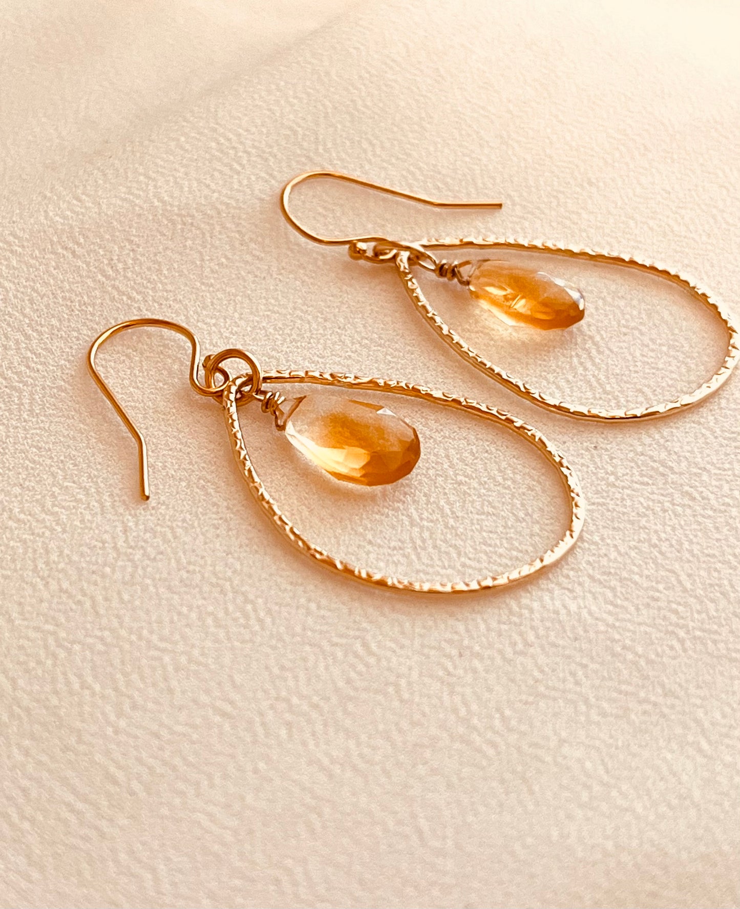 Elegant and lightweight, these handcrafted earrings feature warm yellow teardrop gemstones suspended inside textured 14k gold-filled teardrop hoops. Each stone catches the light with a soft shimmer, adding a refined pop of color to any outfit. The delicate hammered finish on the hoops adds subtle detail and dimension, perfect for both everyday wear and special occasions. Made with care and precision, these earrings combine timeless design with artisan craftsmanship.