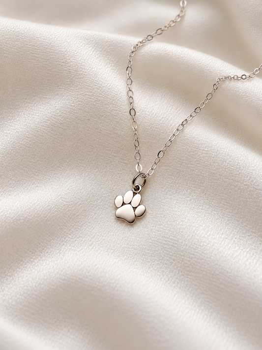 Dainty sterling silver paw print pendant necklace with delicate chain, photographed on soft ivory fabric.