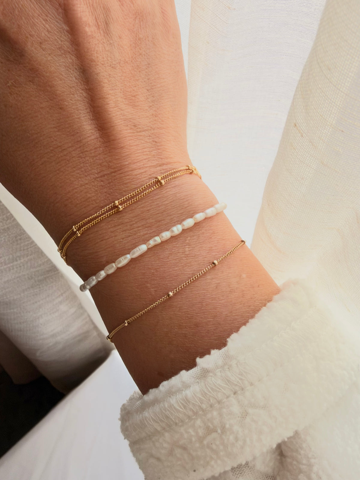 Three delicate bracelets worn on a person's wrist: two thin gold chain bracelets and one white beaded bracelet, styled with a white textured sweater cuff.