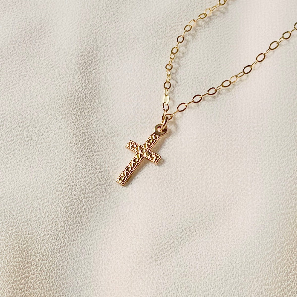 Dainty 14K gold filled cross necklace featuring a tiny 8x15mm double-sided textured cross pendant held against fingers for size reference.